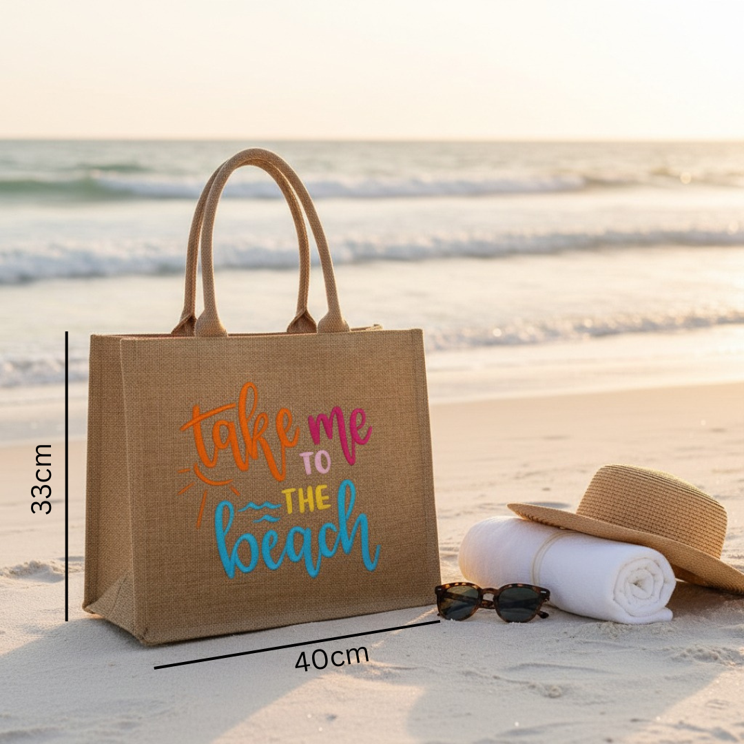 Jute bag with 'Take me to the beach' text on a sandy beach with a hat, sunglasses, and towel.