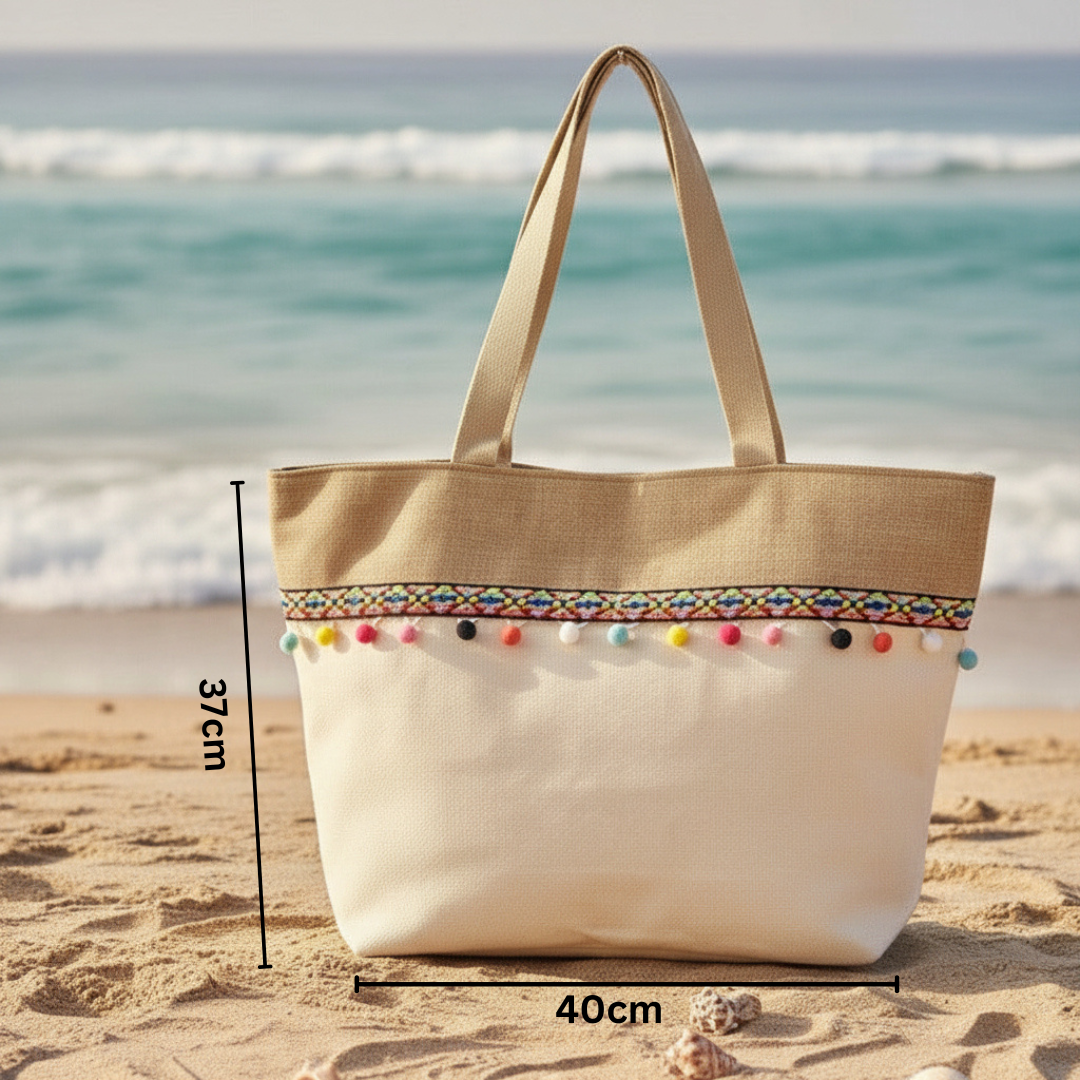 Beige tote bag with colorful pom-poms on a sandy beach with ocean view