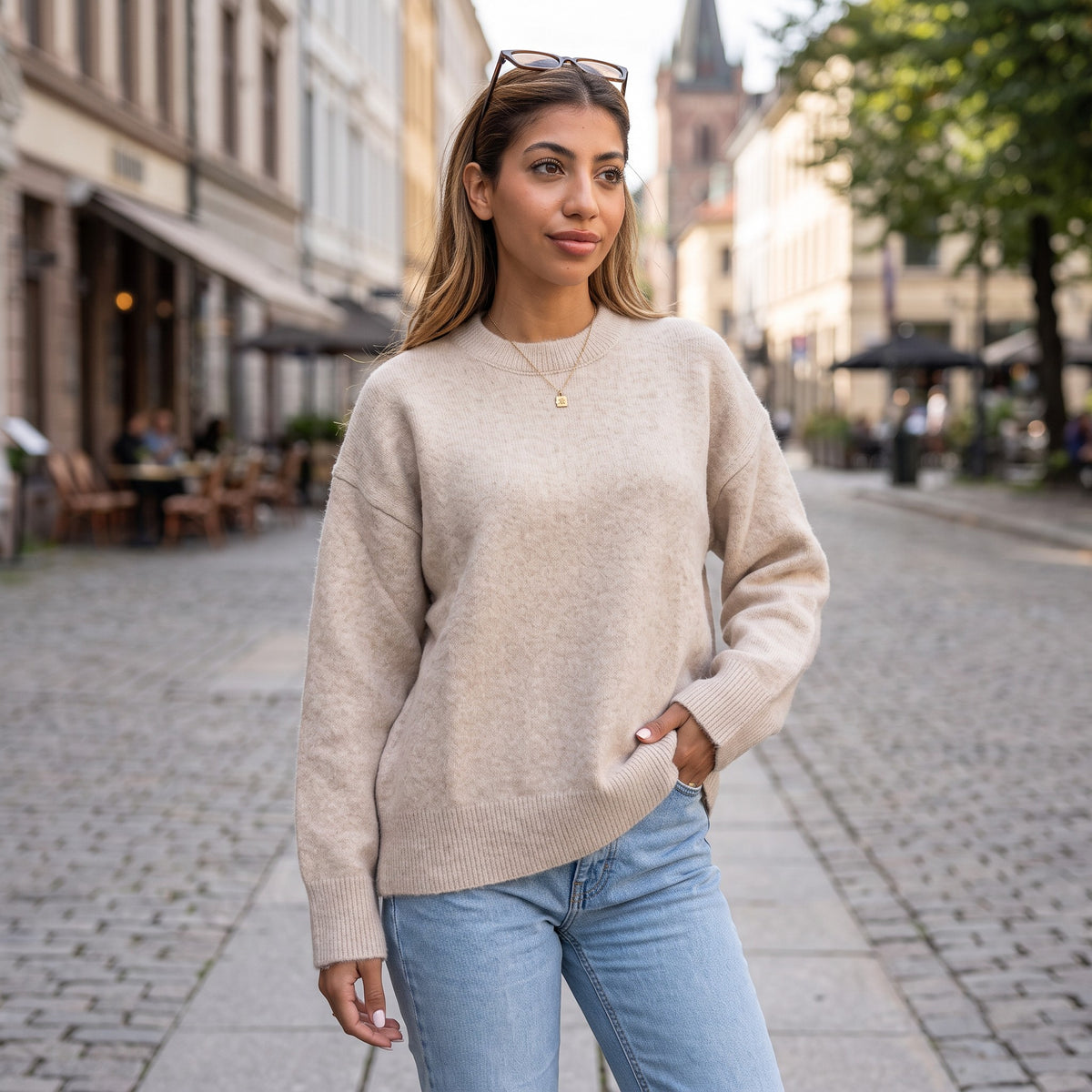 Woman in a beige brushed wool crew neck sweater and light jeans with a gold pendant necklace on a cobbled street