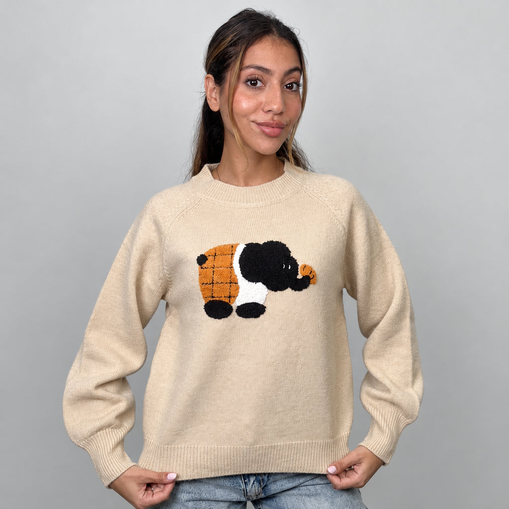Beige knit sweater with a fuzzy elephant patch, holding the ribbed hem