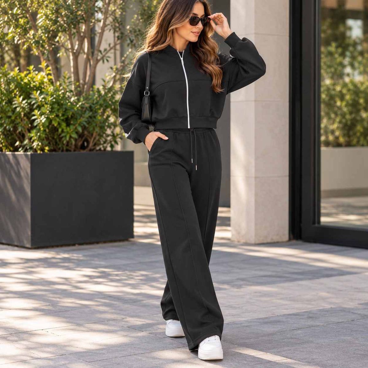 Woman in a black cropped zip jacket and matching wide leg jogging pants with sunglasses and a shoulder bag beside a planter