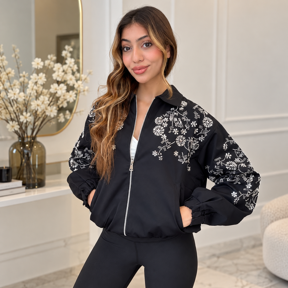 Woman in a black bomber jacket with white beaded flowers on the shoulders and puff sleeves, hands in pockets