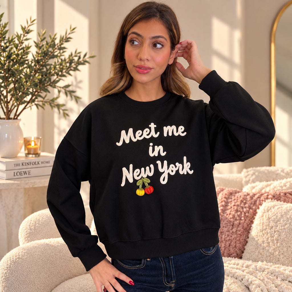 Woman in a black Meet me in New York sweatshirt with cream lettering and cherries, hand at her ear