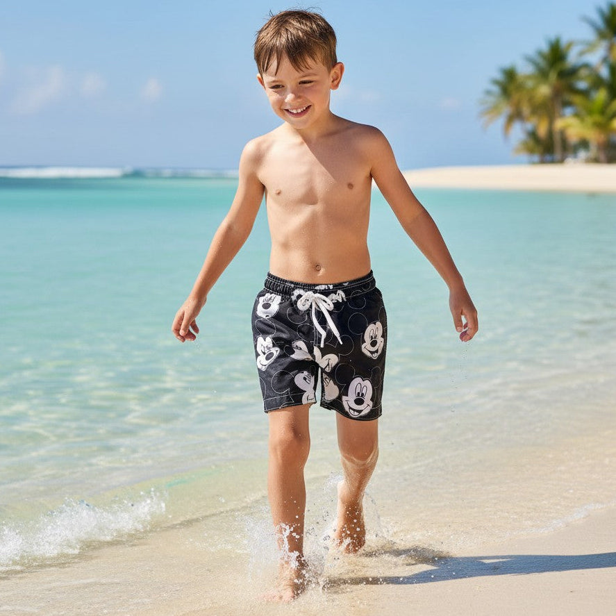 Young boy in black mickey swim shorts walking on a sandy beach with clear blue water