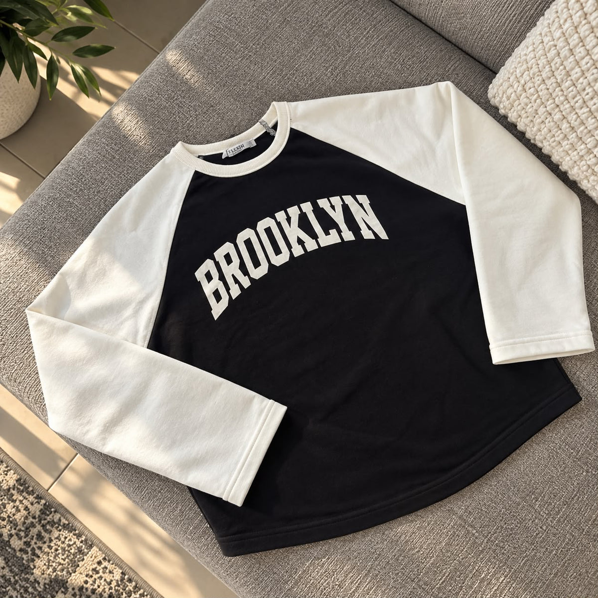Black Brooklyn sweatshirt with white raglan sleeves laid flat on a grey sofa