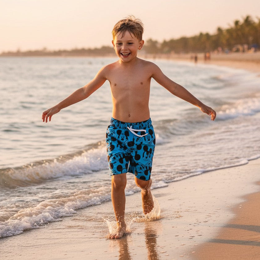 Child in blue swim trunks standing on a beach with waves and palm trees in the background