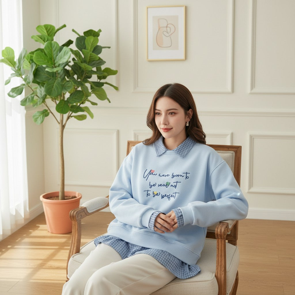 Seated in a wooden armchair wearing a pale blue sweatshirt with checked layers