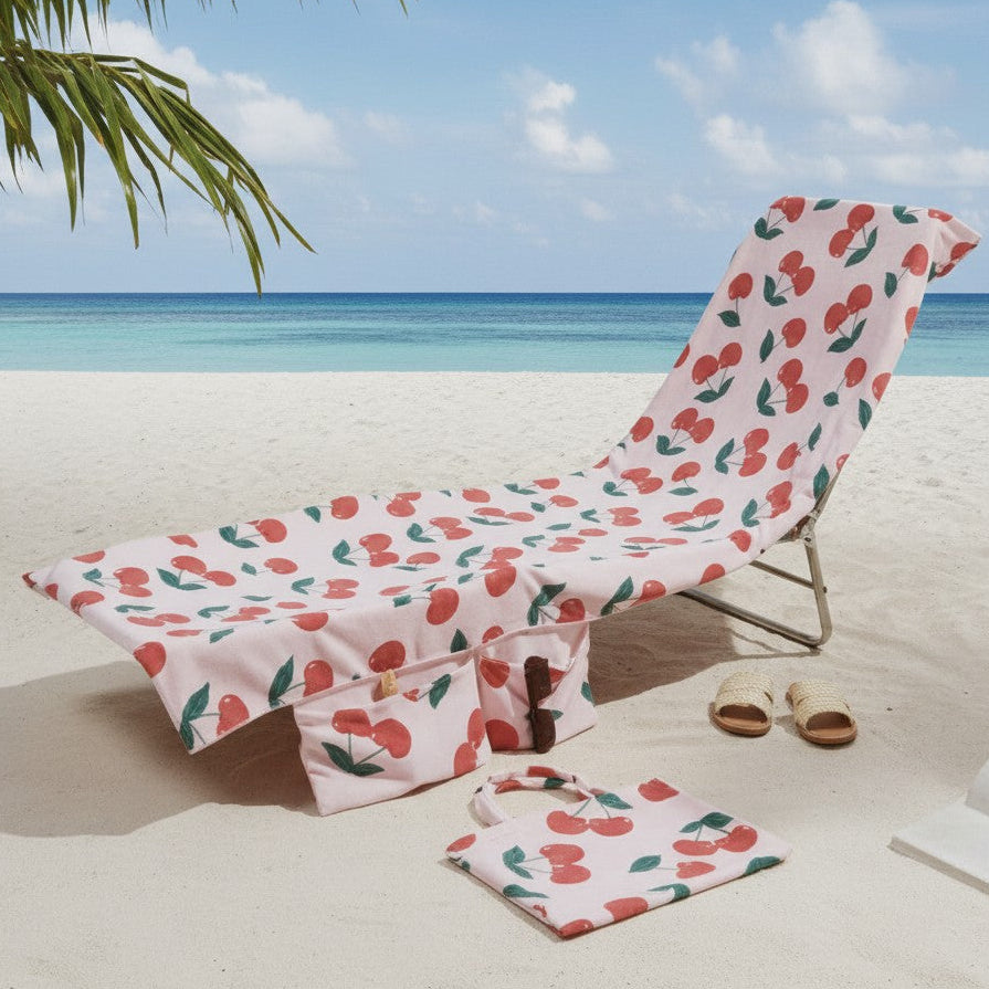 A beach lounge chair and matching towel set with a pink cherry print, placed on white sand at the beach with ocean, sky, and palm leaves visible.