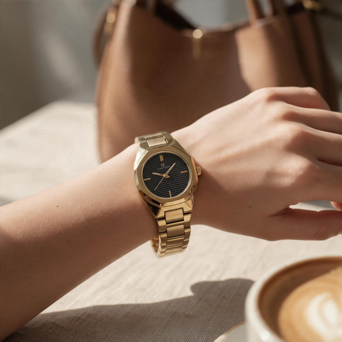 Gold watch with black dial worn on the wrist beside a latte