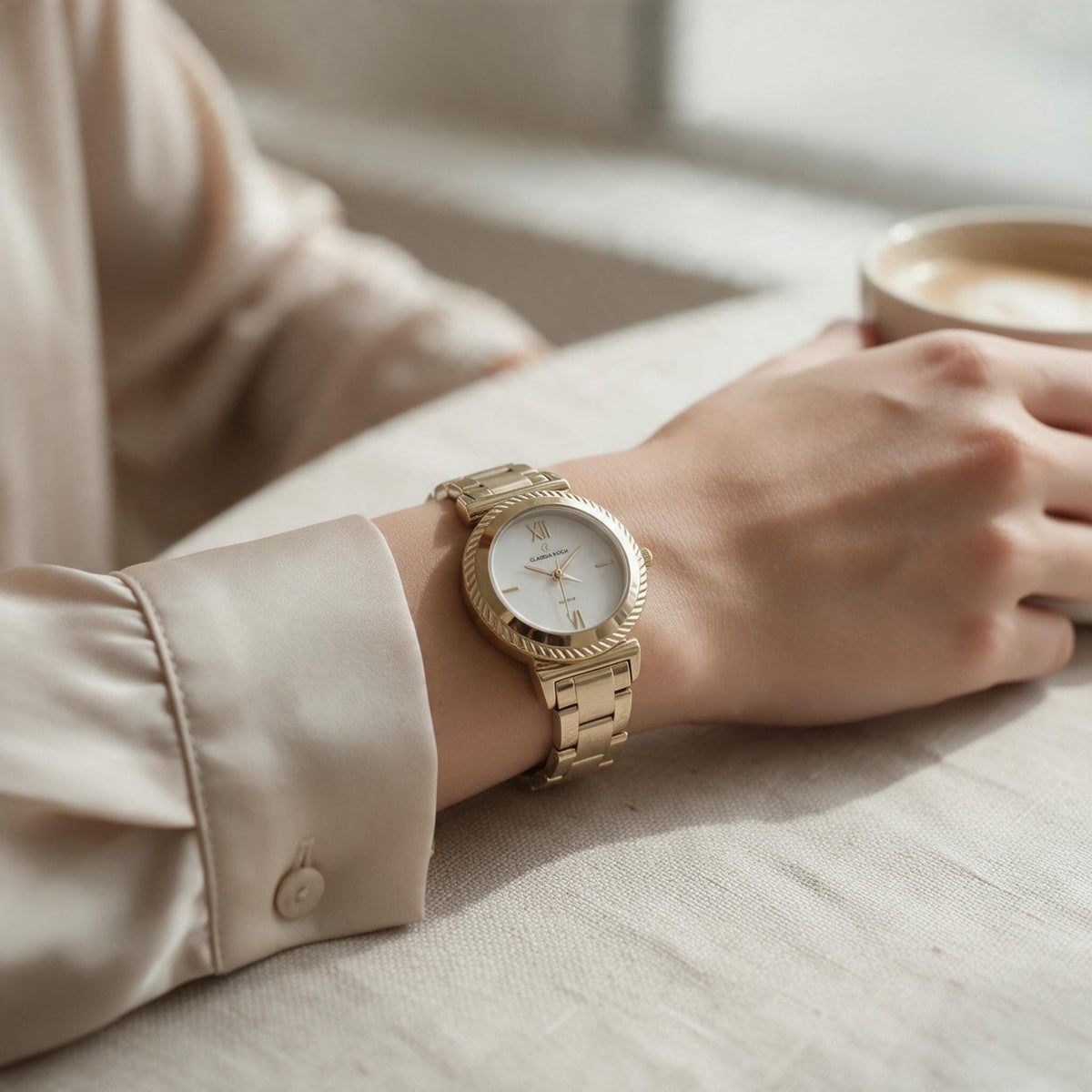 Gold watch with white dial and ridged bezel worn with a cream blouse at a table