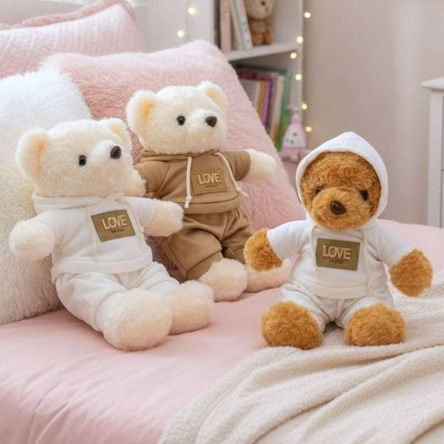 Three tracksuit teddy bears with gold Love chest patches arranged on pink bedding