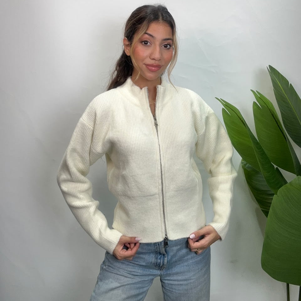 Woman in a cream zip up knit cardigan with hands on her hips against a white wall