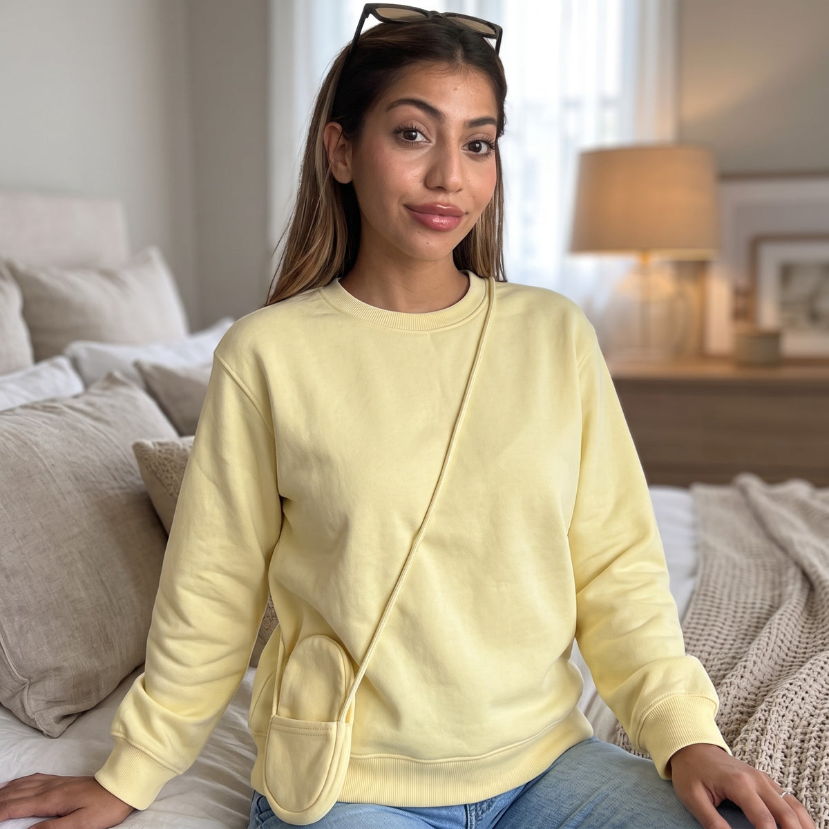 Seated on a bed in a soft yellow sweatshirt, thin strap of the pouch across her body