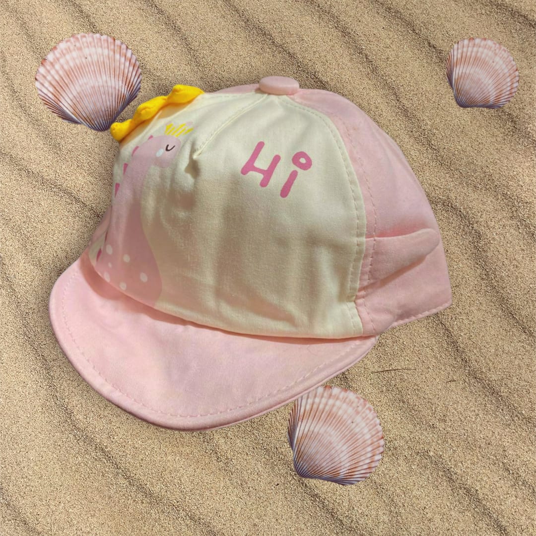 Pale pink and cream infant cap with pink dinosaur artwork and yellow crest spikes, styled beside seashells
