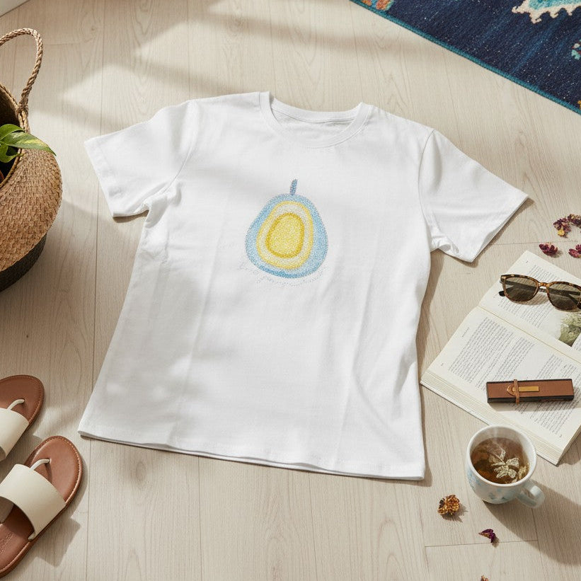 A white short-sleeve T-shirt laid flat on a light wood floor, featuring a colorful avocado graphic on the chest. The shirt is styled in a cozy room setting with a potted plant, slide sandals, sunglasses on an open book, a cup of tea, and a patterned rug nearby.
