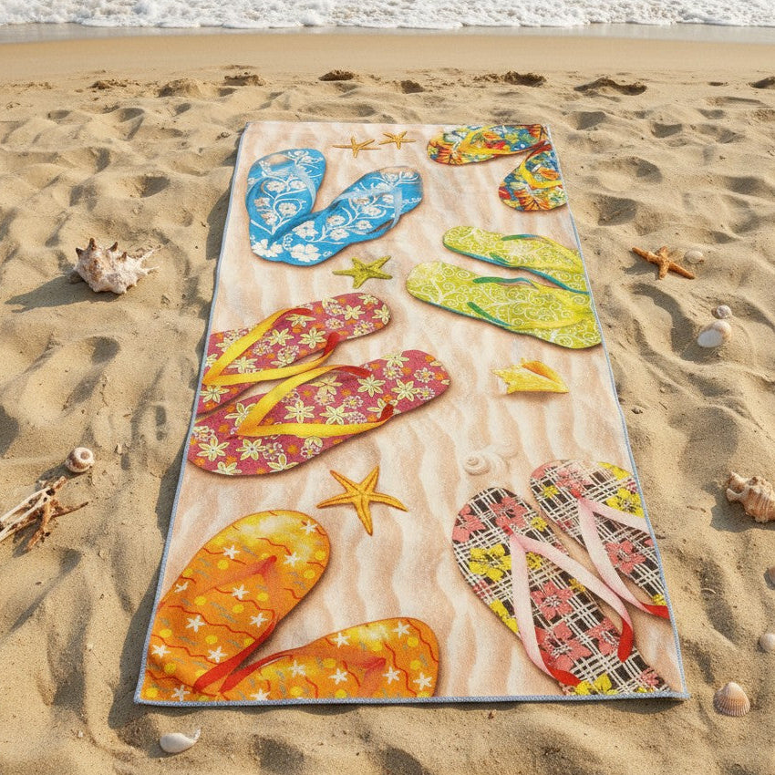 A rectangular beach towel laid flat on sandy shore near the ocean, printed with colorful flip-flop sandals and small starfish motifs. The towel is surrounded by sand, shells, and seaweed, with waves visible in the background.
