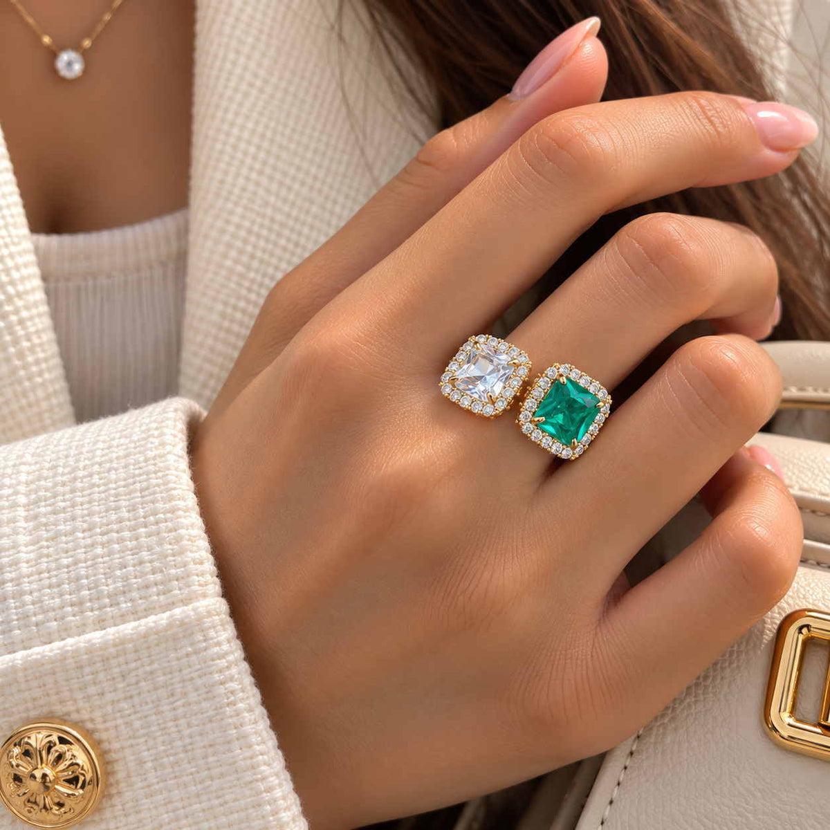 Hand wearing a gold ring with a clear square stone and an emerald green stone, each in a crystal halo