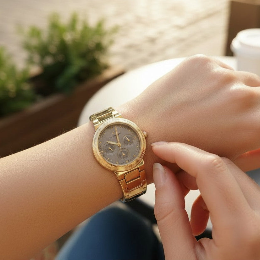 A gold-tone metal link watch with a round dial and matching gold-tone case, worn on a person's wrist with another hand adjusting or touching the strap, in a softly blurred outdoor cafe setting.