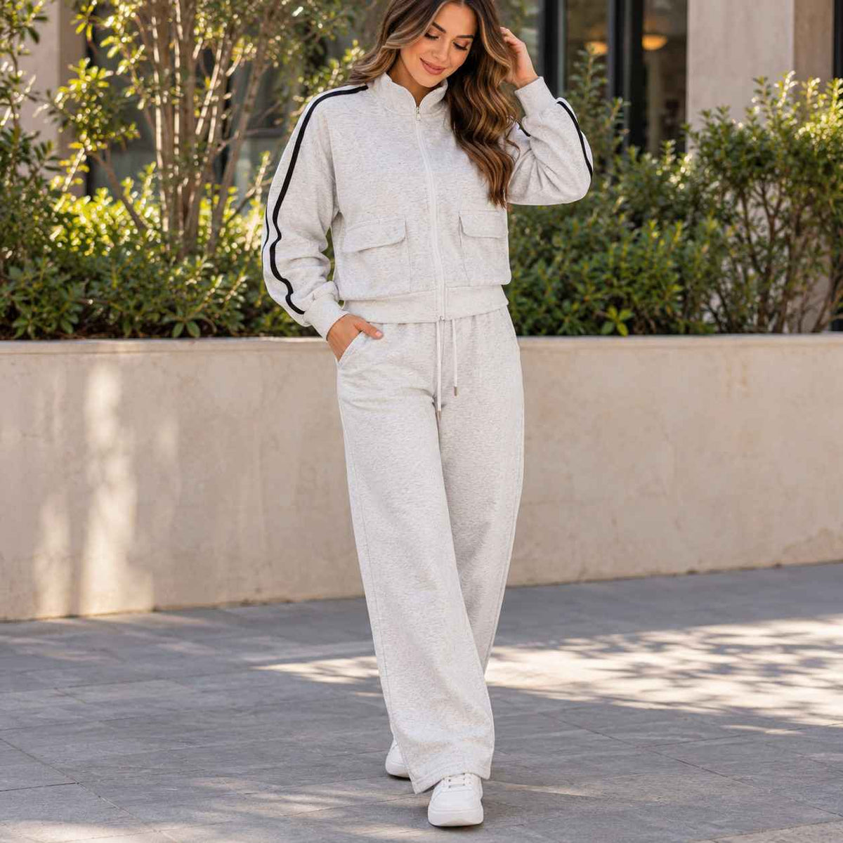 Woman in a grey zip jacket with black sleeve stripes and matching wide leg pants by a stone wall