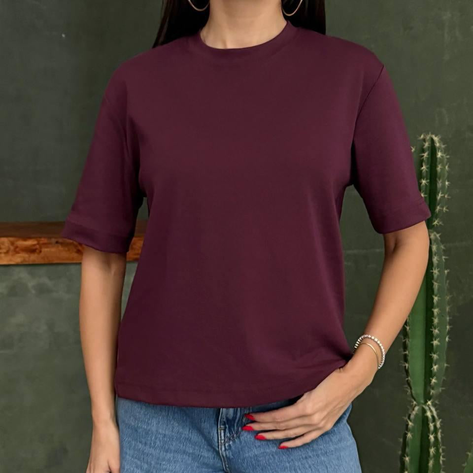 Person wearing a maroon red tshirt and blue jeans standing against a green wall with a cactus plant