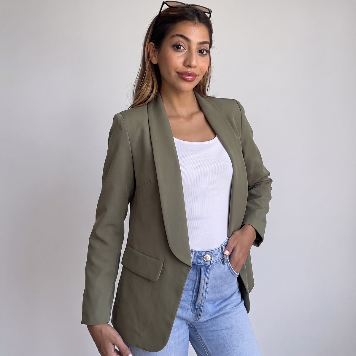 Woman wearing an open olive green shawl collar blazer over a white tank and light jeans