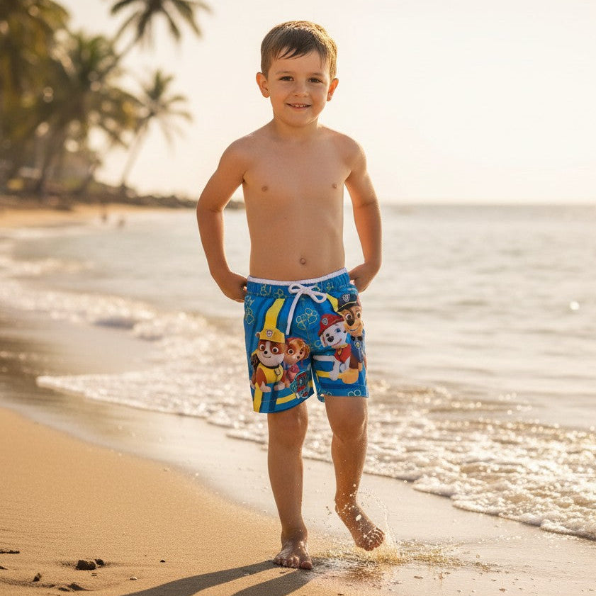 Young boy in colorful swim trunks standing on a sandy beach with palm trees in the background.
