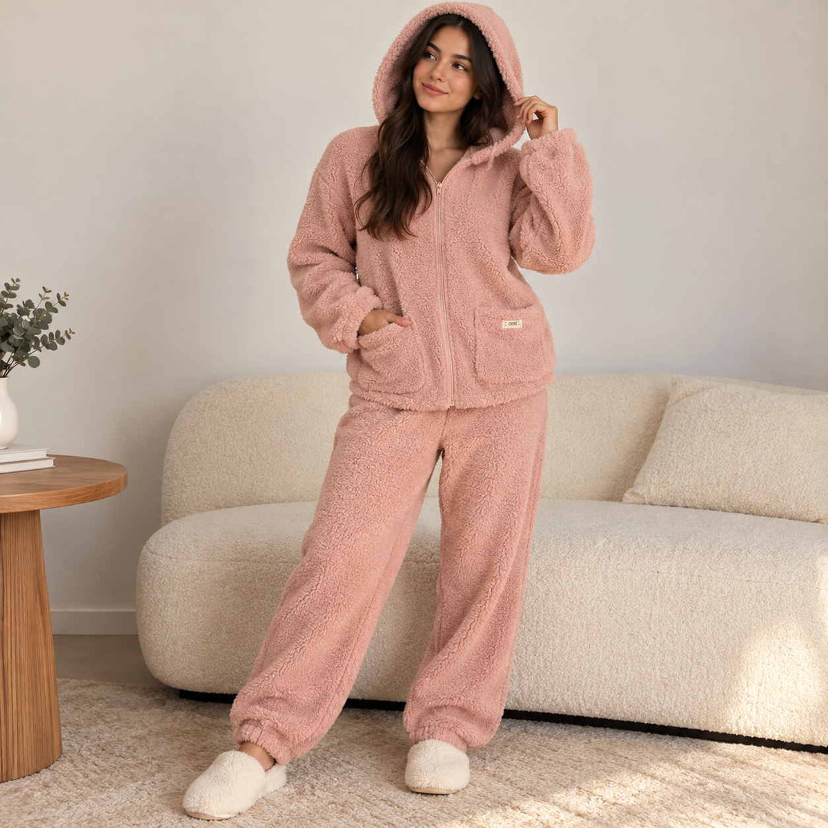 Woman wearing the pink fleece hoodie set with the hood up and fluffy slippers at home