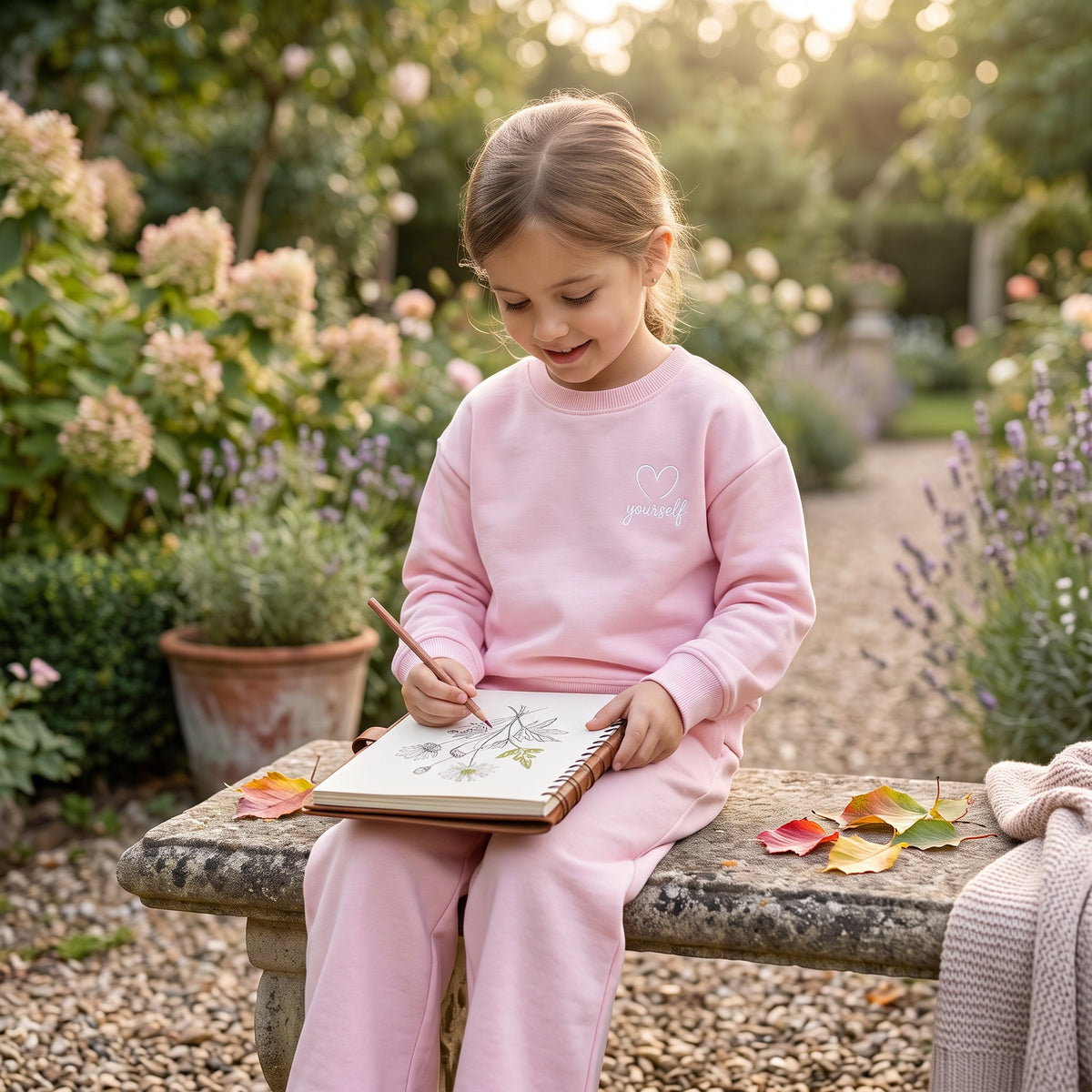 Girl drawing in a sketchbook wearing a pink sweatshirt with white heart yourself chest motif