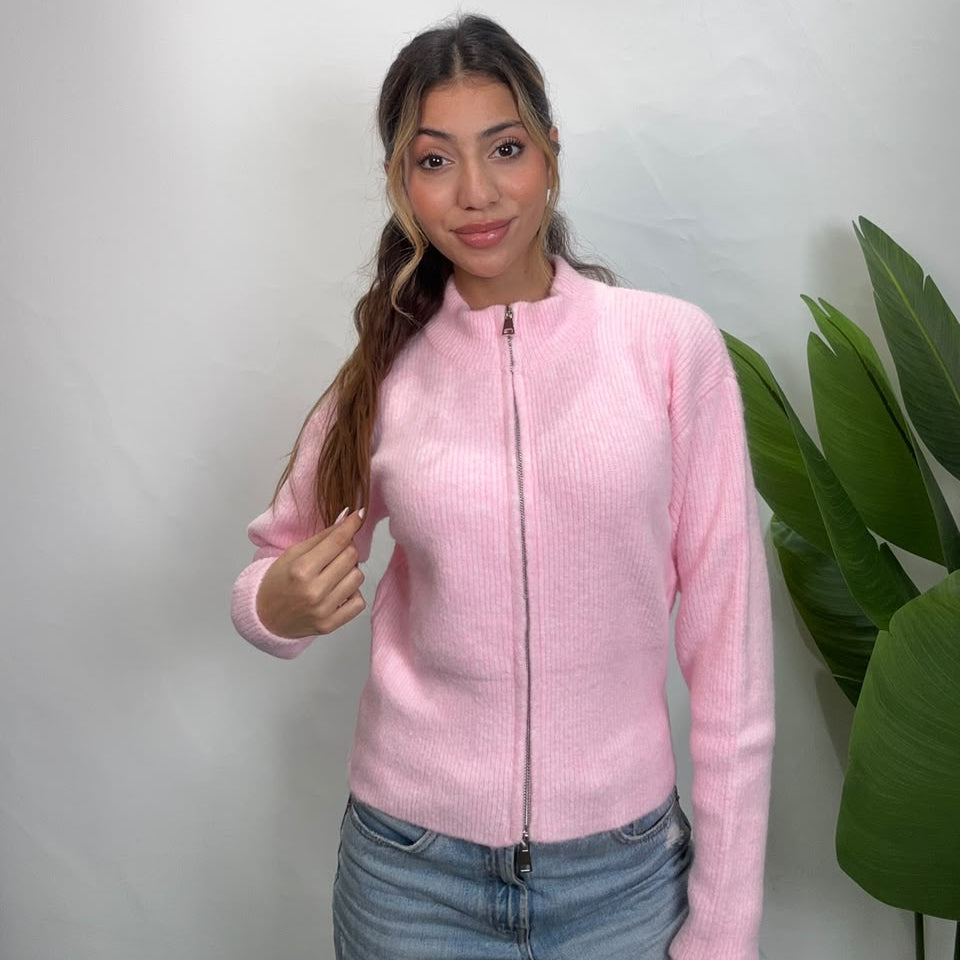 Woman in a baby pink zip up knit cardigan with a funnel neck, holding her ponytail