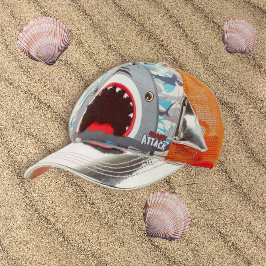 Shark face cap with a white brim and orange mesh back resting on sand beside seashells