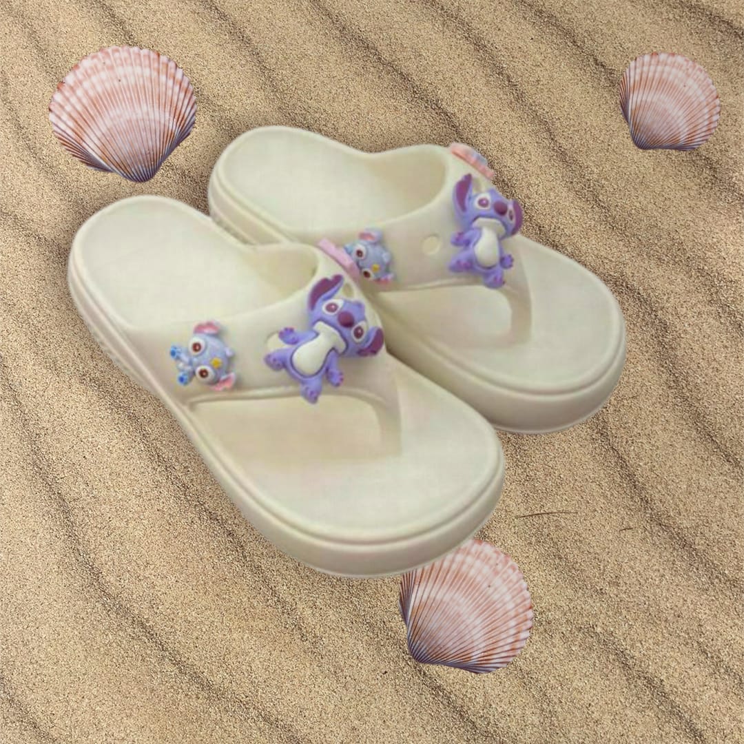 Pair of ivory platform flip flops with lilac charms resting on rippled sand