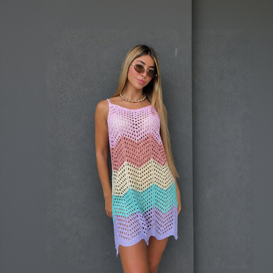 A woman modeling a sleeveless crochet knit mini dress with a pastel chevron pattern in purple, pink, peach, cream, and turquoise, standing against a dark grey textured wall