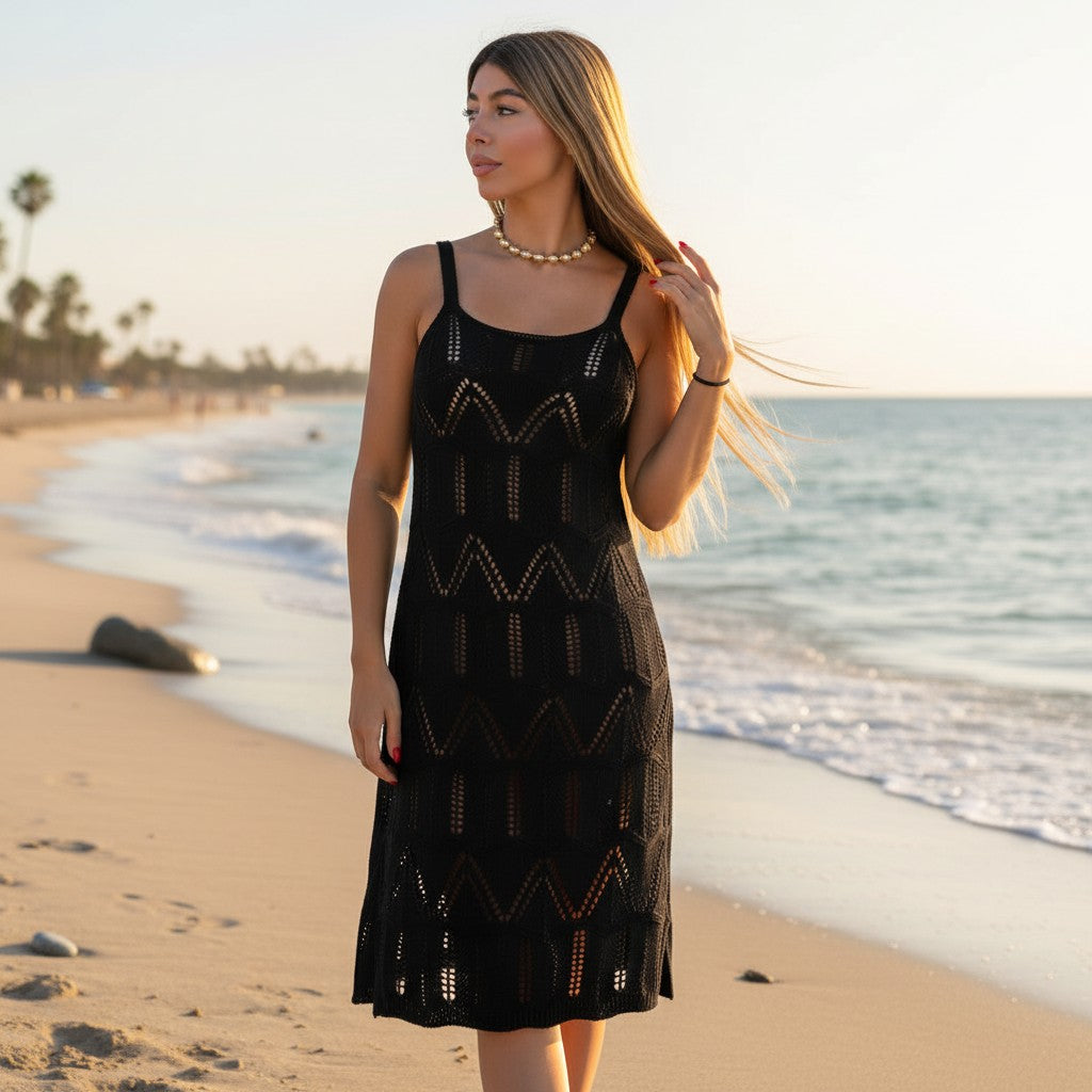 A woman stands on a sandy beach wearing a black sleeveless crochet knit dress with geometric openwork patterns