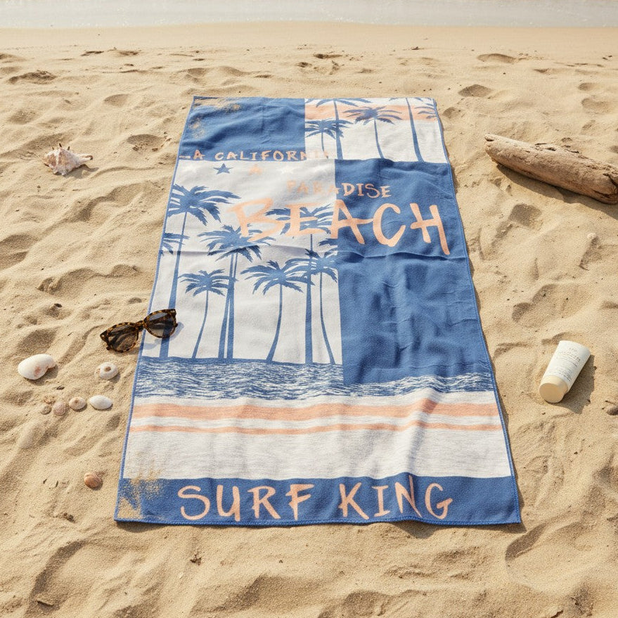 A rectangular beach towel laid flat on a sandy beach near the shoreline, featuring a blue and white tropical palm tree design with the words “A GALLEFORD SURF KING” and “SURF KING.”.