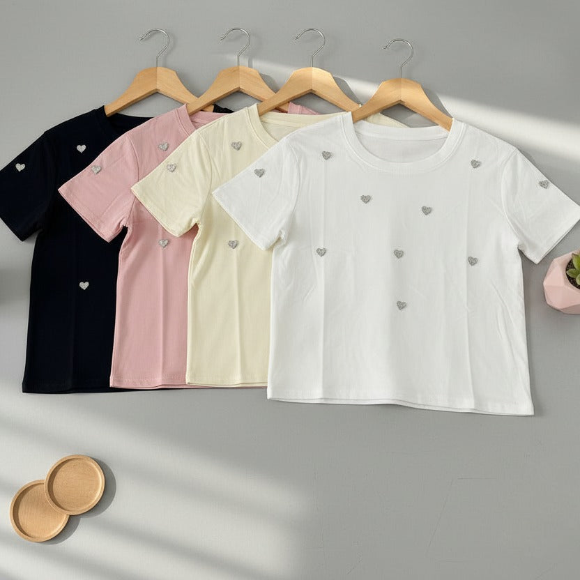 Four t-shirts in black, pink, beige, and white with heart patterns on hangers against a gray background.