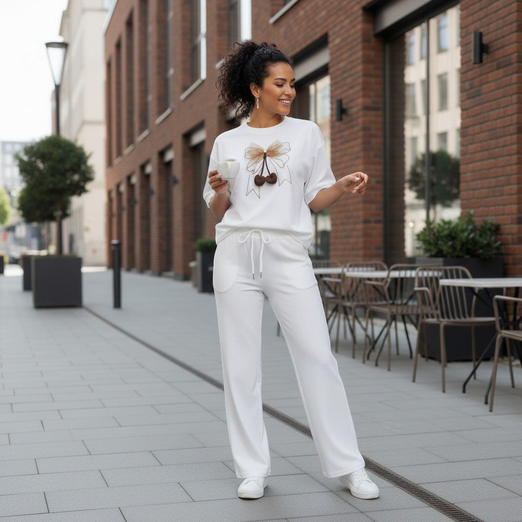 Woman in a white outfit standing on a sidewalk with brick buildings and outdoor furniture in the background.