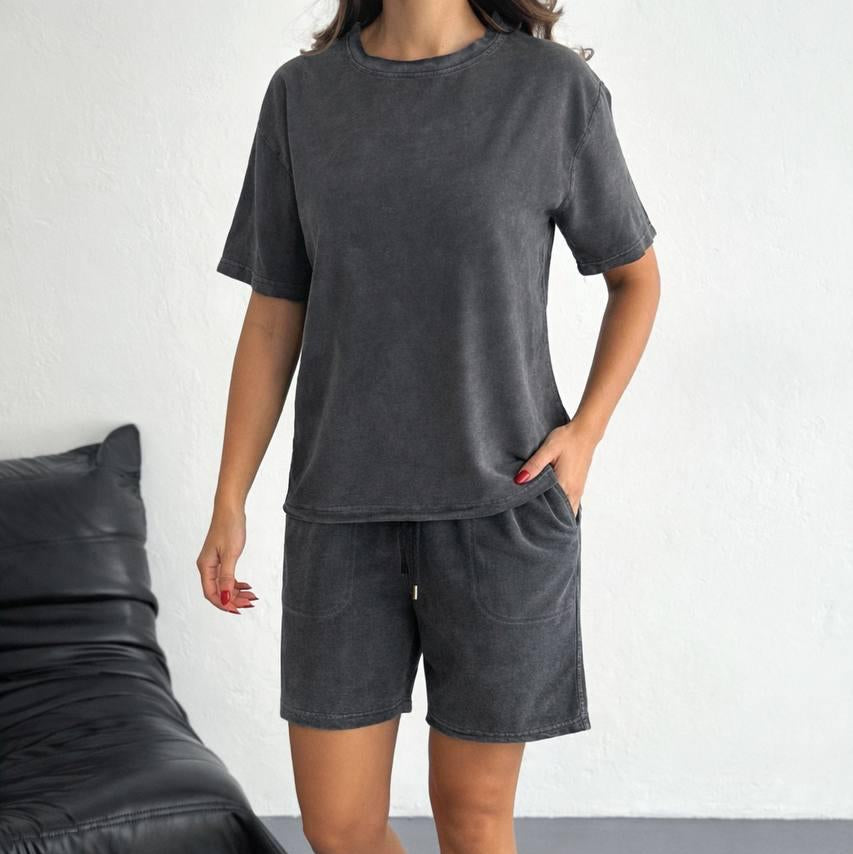 Person wearing a gray t-shirt and shorts set with white sneakers in a minimalistic setting.