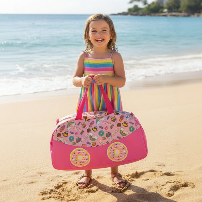 Young girl in a colorful dress holding a pink beach bag with fruit pattern on a sandy beach.