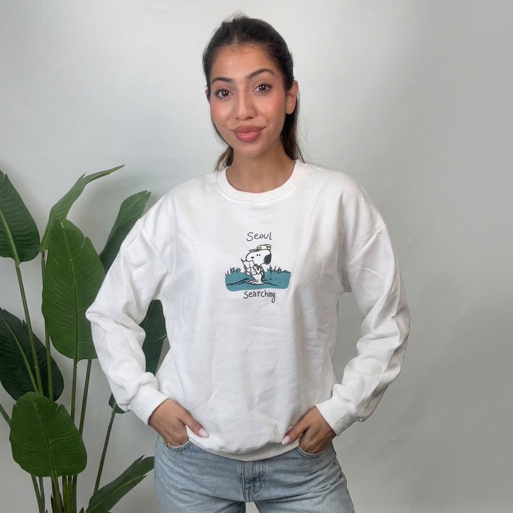 Woman wearing a white crew neck sweatshirt with a teal puppy scout graphic and light jeans