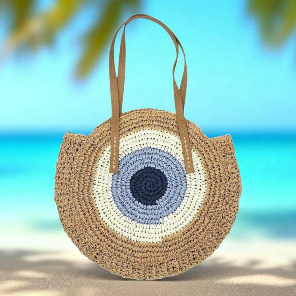 Round woven bag with a blue and white center on a beach with turquoise water and palm leaves.