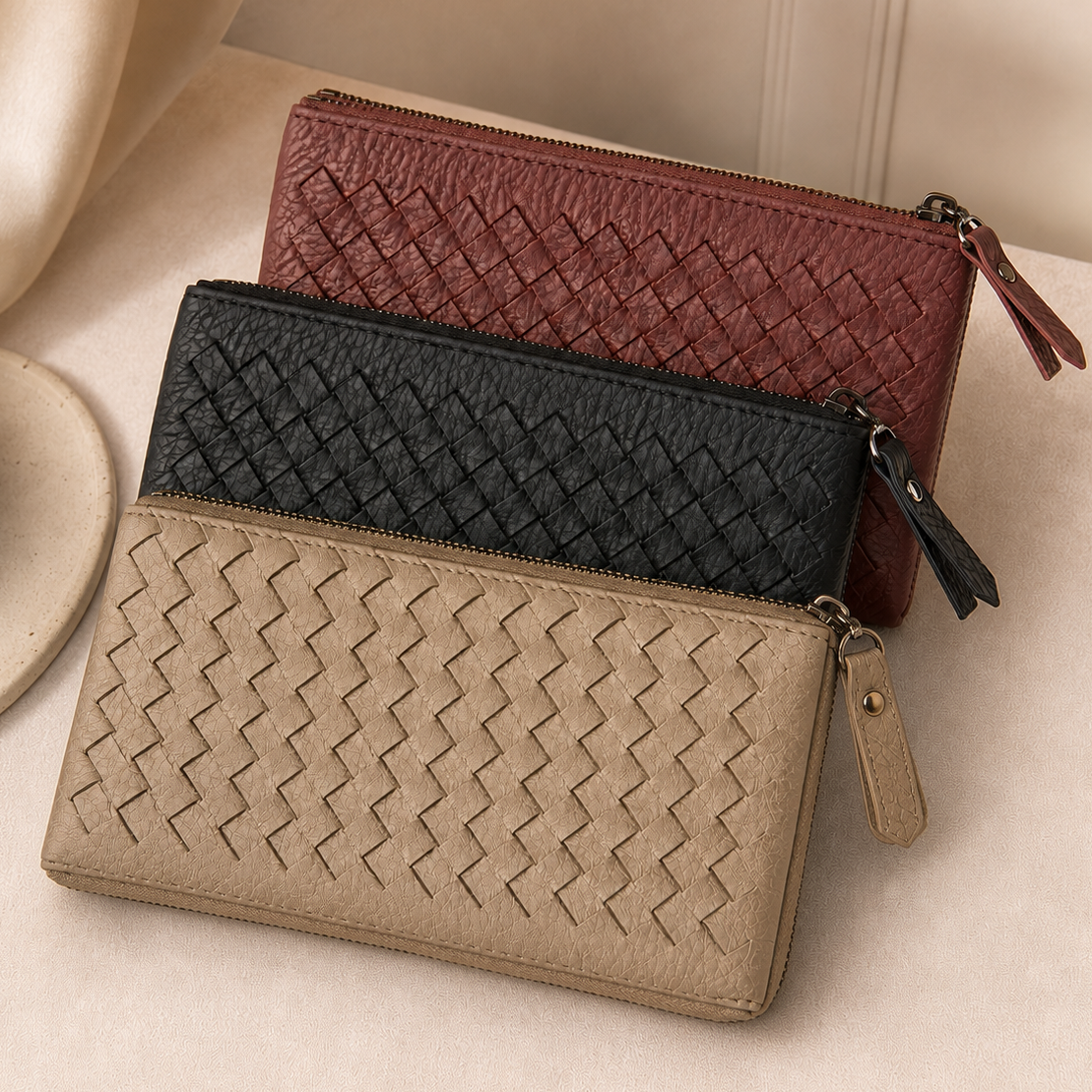 Three woven zip-around wallets in beige, black, and burgundy, arranged diagonally on a soft cream fabric surface.