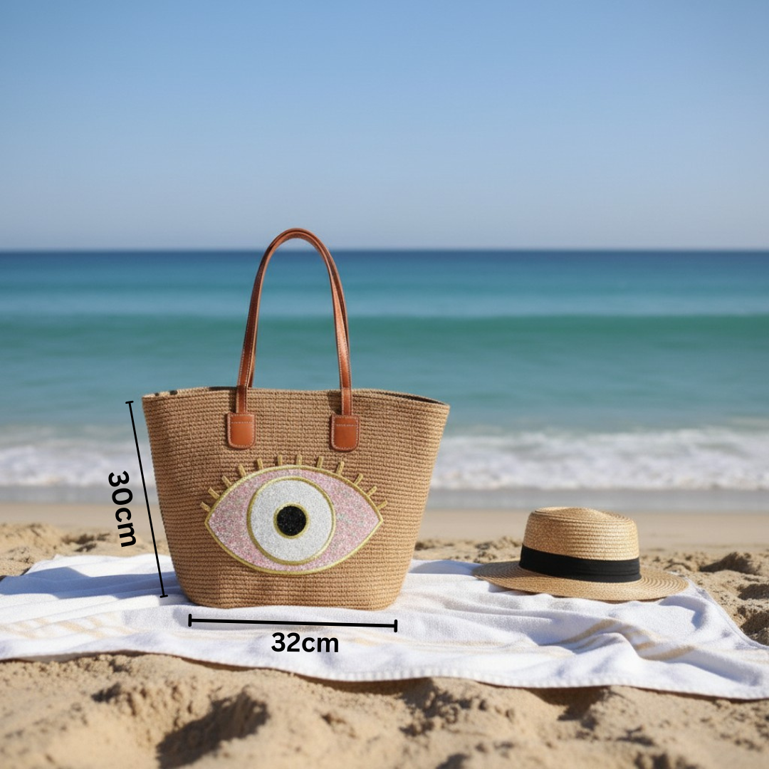 Beige woven bag with eye design and straw hat on a beach with ocean view