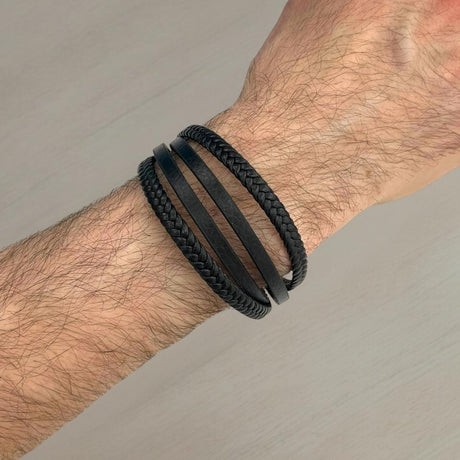 Hand wearing a black 4-strand braided leather bracelet on a neutral background