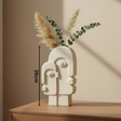 Abstract face-shaped vase with dried plants on a wooden surface against a beige wall.