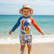 Child in a colorful swimsuit and hat with different animal prints standing on a beach with ocean in the background
