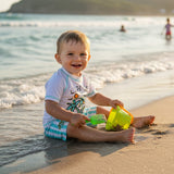 A smiling toddler wearing a white short-sleeve swim shirt with a colorful palm tree graphic and blue-and-white striped shorts
