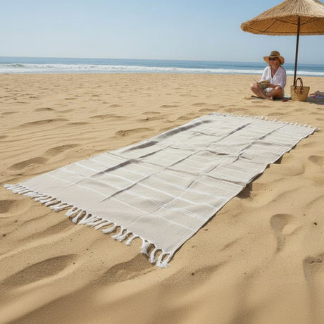 Person sitting under an umbrella on a sandy beach with a towel spread out.
