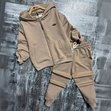 Beige zip up hoodie on a wooden hanger beside matching drawstring joggers on a grey backdrop