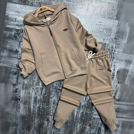Beige zip up hoodie on a wooden hanger beside matching drawstring joggers on a grey backdrop