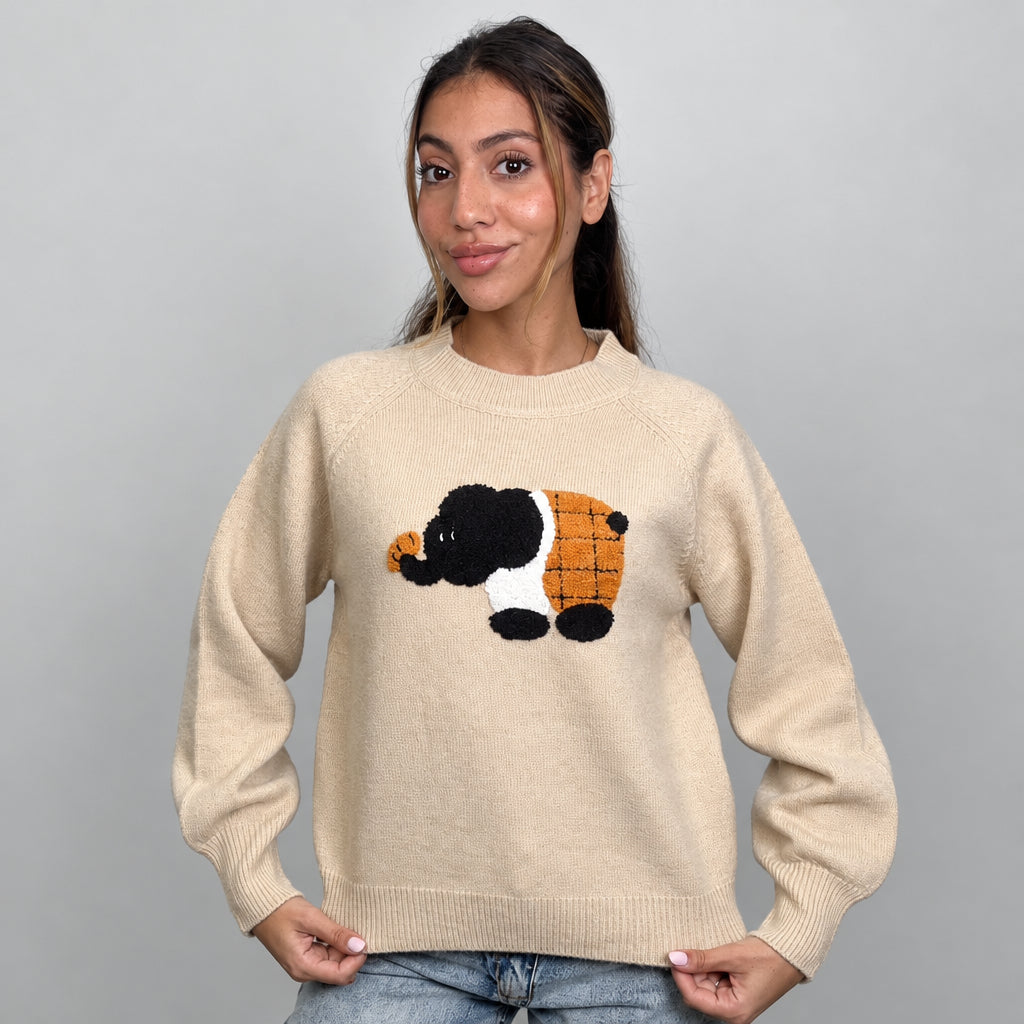 Beige knit sweater with a fuzzy elephant patch, holding the ribbed hem